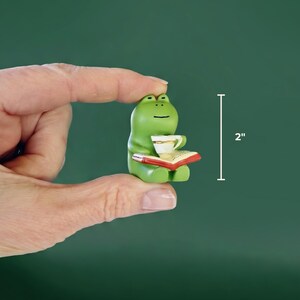 A hand holding a tiny, two-inch tall green ceramic frog figurine reading a book to demonstrate its small, desk-friendly scale against a solid dark green background.