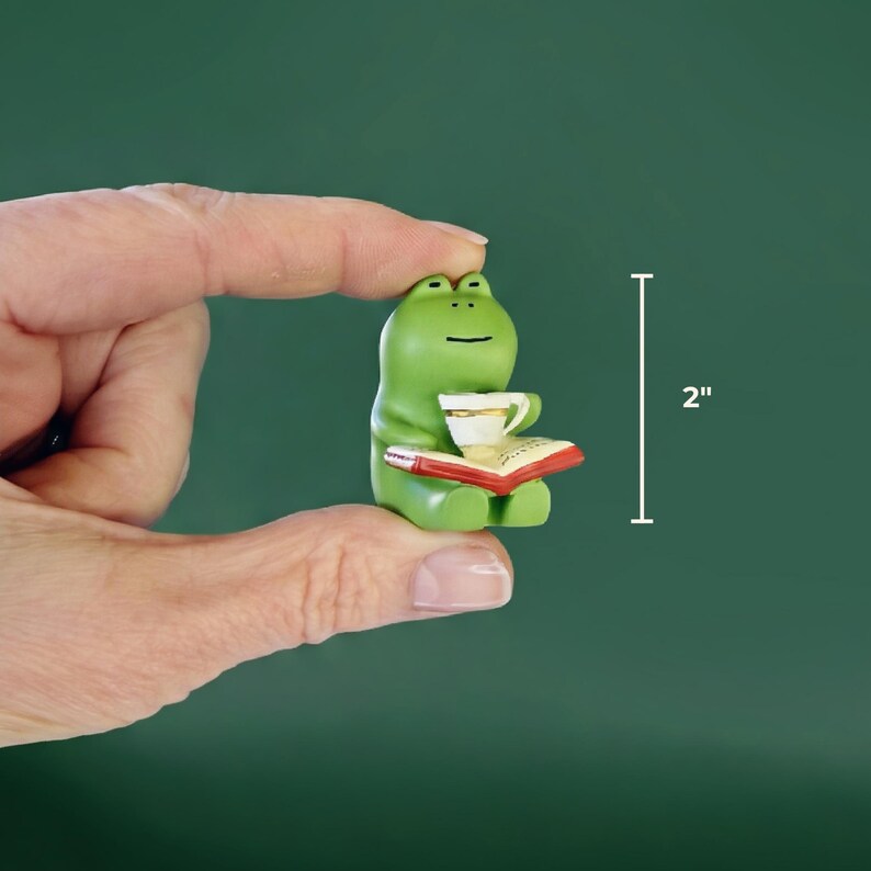 A hand holding a tiny, two-inch tall green ceramic frog figurine reading a book to demonstrate its small, desk-friendly scale against a solid dark green background.