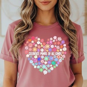 May include: Mauve-colored t-shirt with a heart-shaped floral design. The heart is composed of various colorful flowers and the text "CELEBRATE MINDS OF ALL KINDS" is printed in white. The shirt is paired with blue denim shorts.