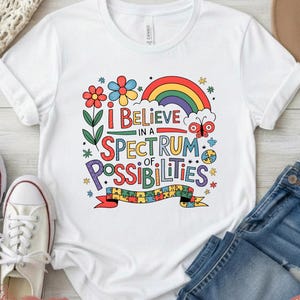 Puede incluir: Camiseta blanca con la frase "I Believe in a Spectrum of Possibilities" en letras coloridas y caprichosas. El diseño incluye un arcoíris, flores y una mariposa. La camiseta se combina con vaqueros azules y zapatillas blancas.