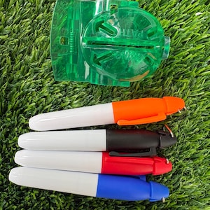 Peut inclure: Ensemble de quatre marqueurs Sharpie orange, noir, rouge et bleu, avec des corps blancs, et un outil d'entraînement au swing de golf vert. Les marqueurs sont posés sur une surface verte.