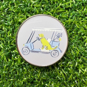 May include: A round golf ball marker with a light gray background and a gold rim. The marker features a yellow Labrador Retriever driving a light blue golf cart. Golf clubs are in the back of the cart.