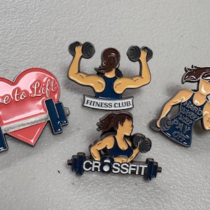 Puede incluir: Colección de pines de esmalte con temas de fitness. Los diseños incluyen un corazón rosa con "Love to Lift", mujeres levantando pesas y las frases "Fitness Club", "CrossFit" y "Hard Work Will Pay Off".