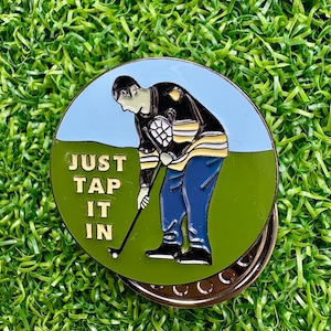 Könnte beinhalten: Runder Golfballmarker mit einem Cartoon-Golfer in blauer Hose und einer schwarz-gelben Jacke, der auf einem grünen Golfplatz puttet. Der Marker trägt den Text "JUST TAP IT IN" in goldenen Buchstaben.