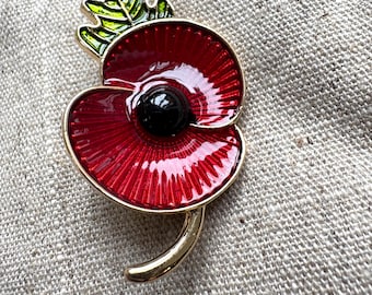 May include: A red enamel poppy brooch with a black centre and gold-tone detailing. The brooch features green and yellow leaves and a curved stem. The poppy is a symbol of remembrance.