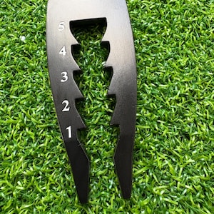 May include: A black golf divot repair tool with a curved top and two prongs. The tool has five numbered slots, from 1 to 5, in white. The tool is designed to repair divots on a golf course and is shown on a green surface.