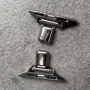以下が含まれることがあります： 2つのステンレススチール製時計バンドアダプター。各アダプターには「STAINLESS STEEL」の文字が刻まれた長方形の留め金が付いています。アダプターはシルバーで、マット仕上げです。背景はテクスチャ加工されたグレーの表面です。