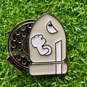 May include: A silver-colored pin featuring a cartoon ghost character with a white outline and a gray body. The pin is set against a backdrop of green artificial turf. The pin has a silver-colored backing.