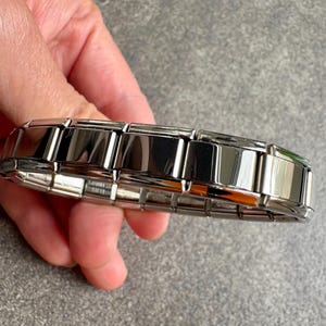 Puede incluir: Una pulsera plateada con un diseño de eslabones rectangulares. Los eslabones están pulidos y son reflectantes, creando una apariencia brillante. La pulsera está sujeta en una mano.