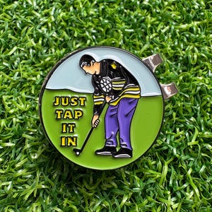 Könnte beinhalten: Runder Golfballmarker mit einer Golfer-Illustration. Der Marker zeigt einen Golfer in schwarz-gelber Kleidung, lila Hose und dem Text "JUST TAP IT IN" auf grünem Hintergrund. Der Marker hat einen silbernen Clip.