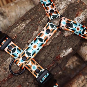 May include: A pet harness featuring a repeating geometric pattern in shades of light blue, orange, black, and white, accented with small evergreen tree motifs. The harness includes black plastic side-release buckles, black metal rivets, and a black D-ring.