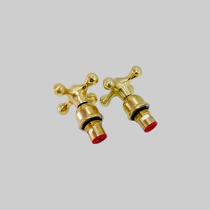 May include: Two brass faucet handles with a cross-shaped design. The handles have a shiny gold finish and red accents at the base. The image is set against a neutral background.