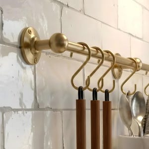 Puede incluir: Un estante dorado para utensilios de cocina montado en una pared de azulejos blancos. El estante tiene una barra horizontal con varios ganchos en forma de S. Utensilios de cocina con mango de madera cuelgan de los ganchos.