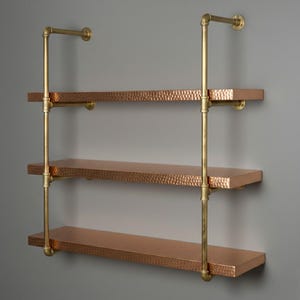 Puede incluir: Estantería de pared de tres niveles con acabado de cobre martillado y tuberías de metal color latón. Los estantes son rectangulares y las tuberías de metal están dispuestas verticalmente. La estantería está montada en una pared gris.