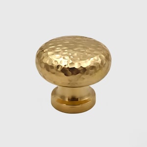 Peut inclure: Un bouton de meuble doré avec une texture martelée. Le bouton a un dessus arrondi et une base cylindrique. La finition est brillante et réfléchissante, adaptée à la quincaillerie de meubles.