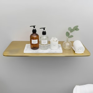 May include: A gold-colored floating shelf displays bathroom essentials. Two amber glass bottles with black pumps, labeled "SAVON MAINS" and "LOTION CORPS", sit on a marble tray. A small white container holds cotton swabs, and a rolled white towel is also present.