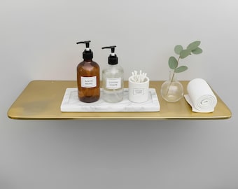 May include: A gold-colored floating shelf displays bathroom essentials. Two amber glass bottles with black pumps, labeled "SAVON MAINS" and "LOTION CORPS", sit on a marble tray. A small white container holds cotton swabs, and a rolled white towel is also present.