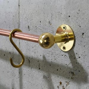 May include: A copper and brass wall-mounted rail with a hook. The rail is made of copper and has a brass ball on one end and a brass hook on the other. It is mounted on a concrete wall.