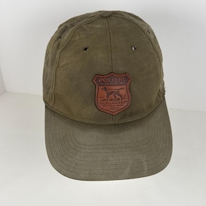 May include: An olive green baseball cap featuring a brown leather shield-shaped patch on the front. The patch displays the text POLO RL GUIDE, RALPH LAUREN & CO, SPORTSMAN APPAREL, 1967, and an illustration of a dog. The cap has metal eyelets.