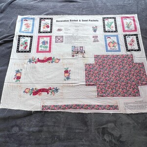 May include: A white fabric panel with a variety of printed designs. The panel features framed images of flowers, vegetables, and fruit, along with floral and ribbon accents. The text "Decorative Basket & Seed Packets" is printed at the top.