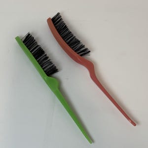 May include: Two hair styling combs. One is green, the other is a reddish-orange. Both have black bristles and long, pointed handles. The combs are designed for sectioning and styling hair. The handles are curved for a comfortable grip.