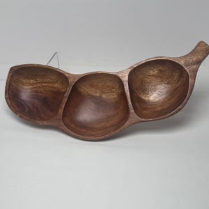 Puede incluir: Una bandeja de madera para servir con tres compartimentos redondeados conectados y un asa. La bandeja está hecha de madera marrón oscuro con vetas visibles. Ideal para servir aperitivos o entremeses.