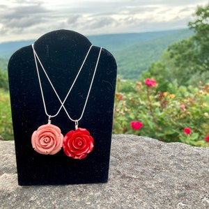 May include: Two silver chain necklaces with rose-shaped pendants. The pendant on the left is a light pink rose, and the pendant on the right is a bright red rose.