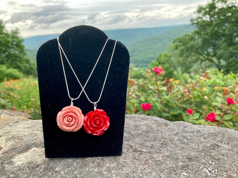 May include: Two silver chain necklaces with rose-shaped pendants. The pendant on the left is a light pink rose, and the pendant on the right is a bright red rose.