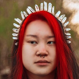 May include: A woman with bright red hair is wearing a crystal crown headband. The crown is made of clear quartz crystals that are wired together. The woman is looking at the camera with a serious expression.