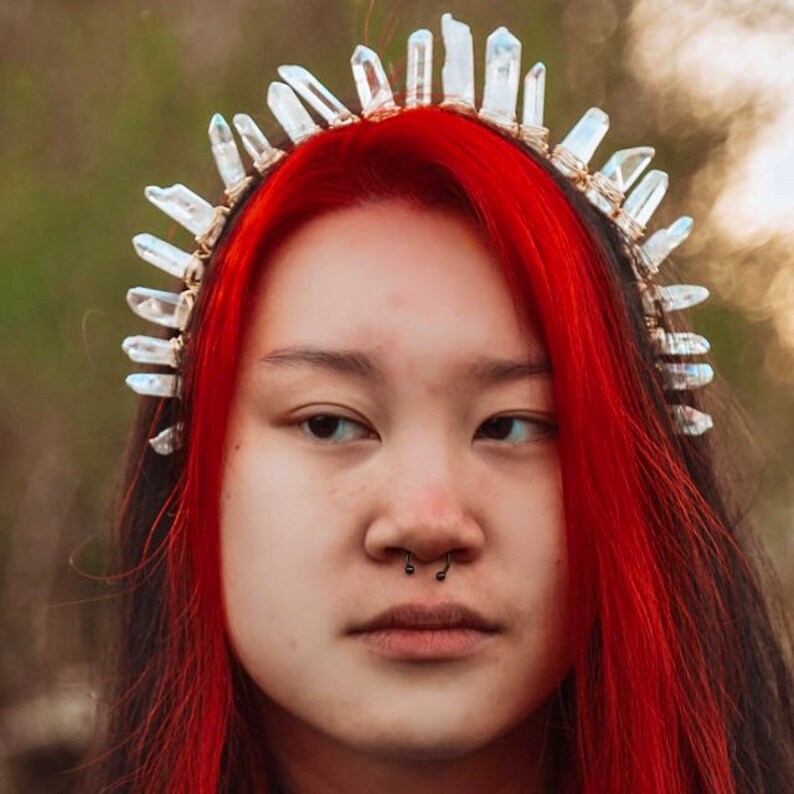 May include: A woman with bright red hair is wearing a crystal crown headband. The crown is made of clear quartz crystals that are wired together. The woman is looking at the camera with a serious expression.