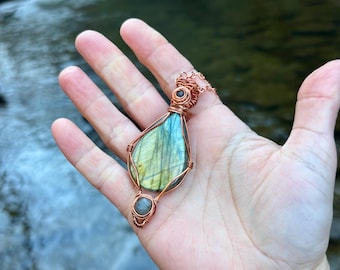 Peut inclure: Un collier pendentif en fil de cuivre avec une grande pierre de labradorite en forme de goutte d'eau aux teintes bleues, vertes et jaunes. Une petite pierre ronde est sertie en dessous. Le collier est tenu dans une main.