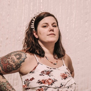 May include: A woman with short brown hair wearing a white floral tank top and a crystal crown headband. She has a large tattoo on her upper arm of a woman sitting in a rose.
