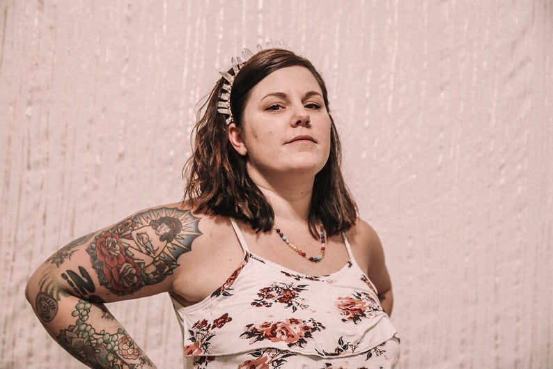 May include: A woman with short brown hair wearing a white floral tank top and a crystal crown headband. She has a large tattoo on her upper arm of a woman sitting in a rose.