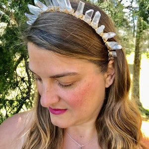 May include: A crystal headband with clear quartz points wrapped in gold wire. The headband is worn by a person with long blonde hair and a purple crystal pendant necklace.