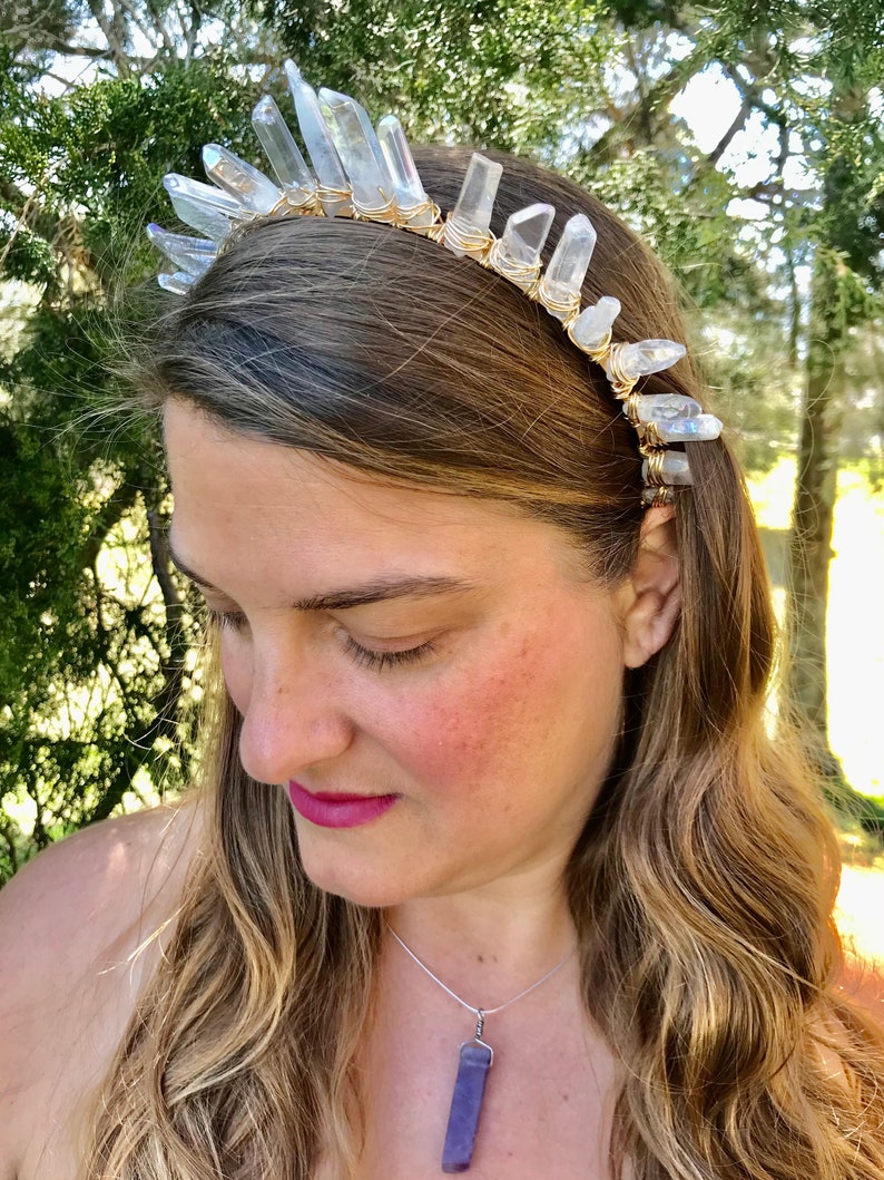 May include: A crystal headband with clear quartz points wrapped in gold wire. The headband is worn by a person with long blonde hair and a purple crystal pendant necklace.
