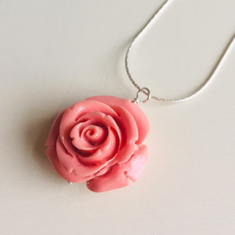 May include: A delicate silver chain necklace with a pink rose pendant. The rose is carved in detail and has a glossy finish.