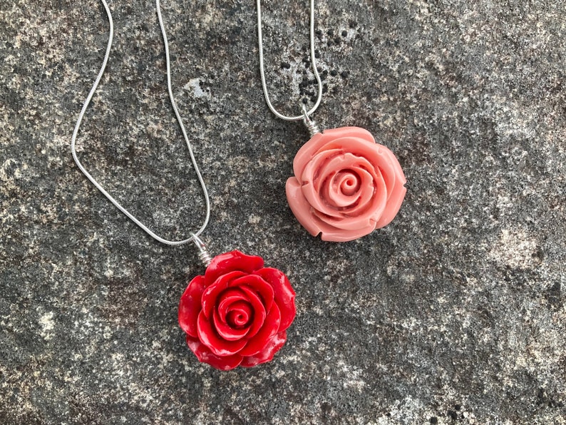 May include: Two silver chain necklaces with rose-shaped pendants. One pendant is a light pink rose, the other is a bright red rose.