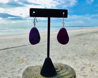Könnte beinhalten: Ein Paar Hängeohrringe mit violetten Achat-Steinen und silbernem Draht, der um die Steine gewickelt ist. Die Ohrringe hängen an einem schwarzen Ohrringständer.