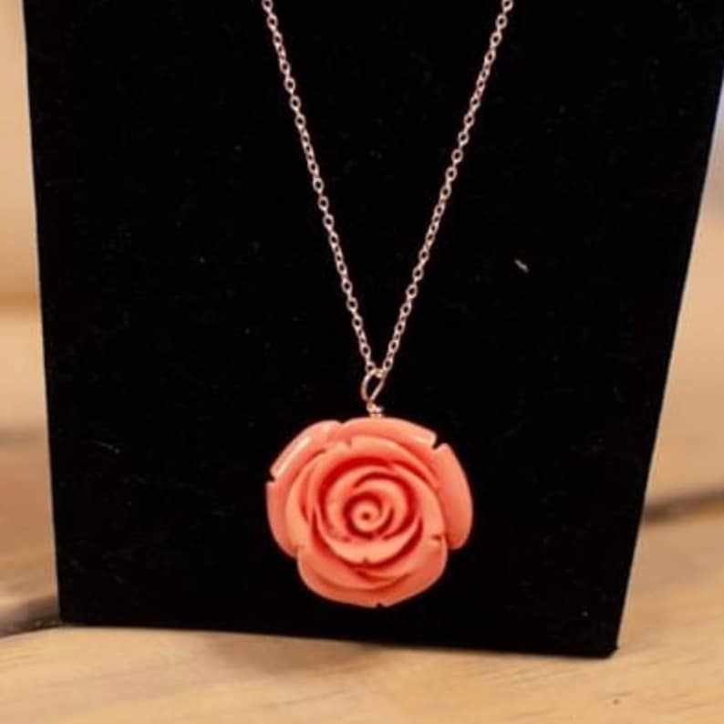 May include: A delicate silver chain necklace with a pink coral rose pendant. The rose is carved with intricate details and has a natural, organic look.