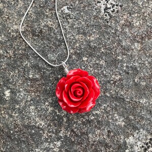 May include: A silver chain necklace with a red rose pendant. The rose is made of a smooth, shiny material and has a detailed design.