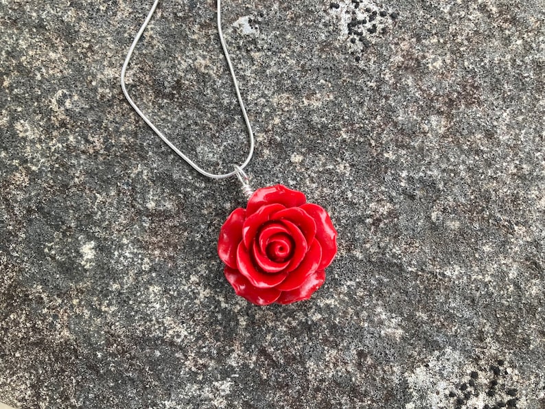 May include: A silver chain necklace with a red rose pendant. The rose is made of a smooth, shiny material and has a detailed design.
