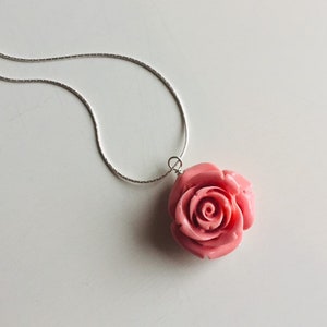 May include: A silver chain necklace with a pink rose pendant. The rose is carved and has a detailed design.