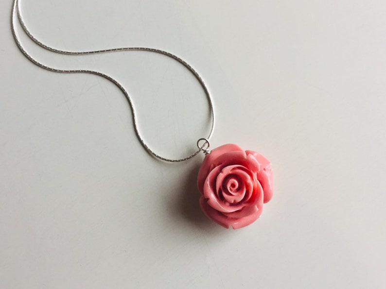 May include: A silver chain necklace with a pink rose pendant. The rose is carved and has a detailed design.