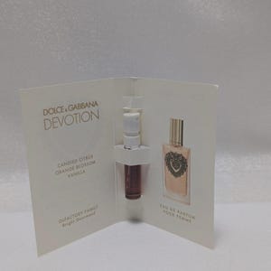 May include: A white card with a perfume sample and a miniature perfume bottle. The card displays "DOLCE & GABBANA DEVOTION" and lists fragrance notes: "CANDIED CITRUS ORANGE BLOSSOM VANILLA". The bottle is labelled "EAU DE PARFUM POUR FEMME".