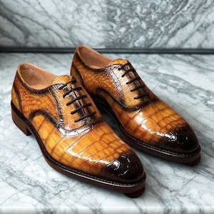 Può includere: Un paio di scarpe eleganti in pelle marrone con una texture a pelle di coccodrillo. Le scarpe hanno lacci marrone scuro e una finitura lucida, con un puntale e suola marrone più scuro. Sono esposte su una superficie di marmo.