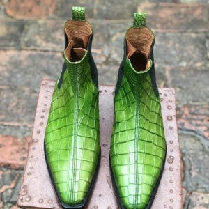 May include: A pair of bright green Chelsea boots featuring a textured crocodile skin pattern, a pointed toe, black elastic side panels, and a small pull tab at the top of the heel collar. The interior lining is a light tan leather material.
