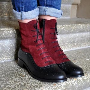 May include: Ankle boots featuring a deep red suede upper and a black leather lower section with brogue detailing. The boots have a front lace-up closure with thin black laces and a side zip for fastening, set on a low heel with a rounded toe.