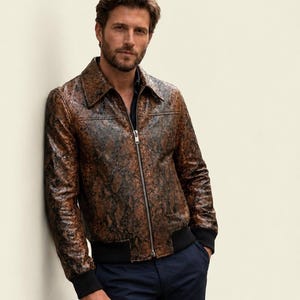 Può includere: Una giacca in pelle con motivo a pelle di serpente marrone e nero, dotata di colletto a punta, cerniera frontale argentata a tutta lunghezza e polsini neri a coste sulle maniche e sull'orlo, abbinata a pantaloni blu scuro e camicia scura.
