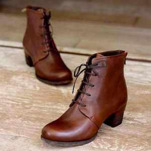 Peut inclure: Une paire de bottines en cuir marron avec un bout arrondi, un laçage frontal avec des lacets marron foncé et un talon bloc empilé. Les bottines présentent une finition lisse et une fermeture éclair à l'arrière, offrant un aspect structuré.
