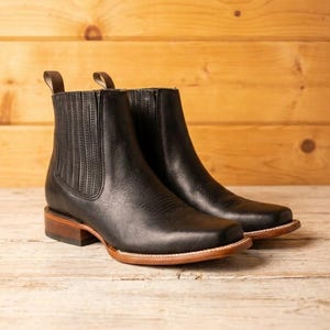 Könnte beinhalten: Ein Paar schwarze Chelsea-Stiefel aus Leder mit quadratischer Zehenpartie, elastischen Seiteneinsätzen mit vertikalen Nähten, einem braunen Absatz in Schichtoptik und einem hellbraunen Rahmen. Jeder Stiefel hat eine braune Zuglasche am Schaft.